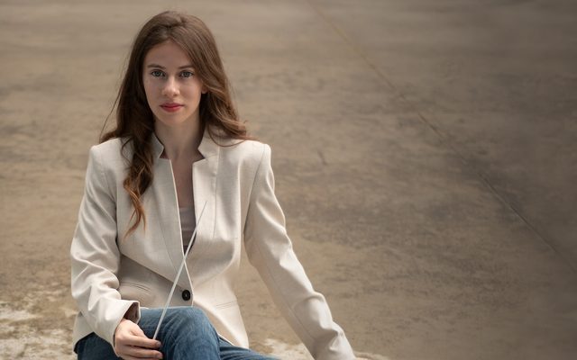 sitting on a rough concrete floor, a young woman in a white jacket and jeans holds a conductor's baton