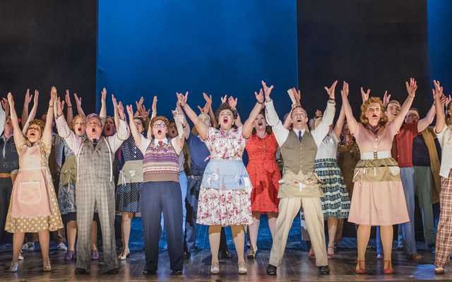 A group stand up-lit and singing passionately with arms upraised in Opera North's production of Kiss Me Kate