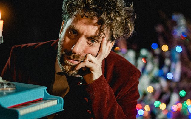 A miserable looking man with a beard, flanked by a toy piano and candle in the foreground and a Christmas tree with lights in the background