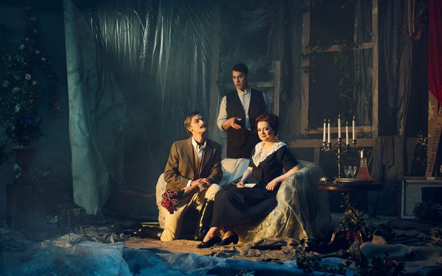 An aristocratic looking man sits on a elegant sofa offering a bunch of flowers to a woman who sits smiling next to him. A man, Figaro, stands over them pointing and looking confused for Opera North's production of The Marriage of Figaro