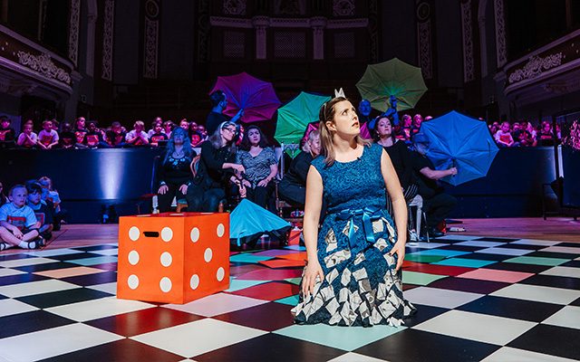 A woman in a blue dress kneeling on the floor beside a big red dice.