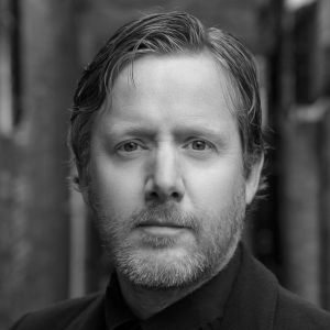 Nicholas Watts has a short beard and is wearing a dark suit.